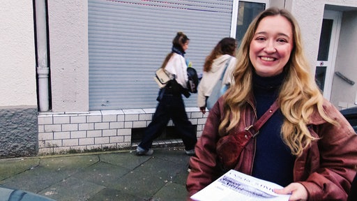 Eine junge Frau steht auf der Straße und lacht in die Kamera, sie hält einen Stapel Flugblätter in der Hand, im Hintergrund gehen Menschen vorbei.