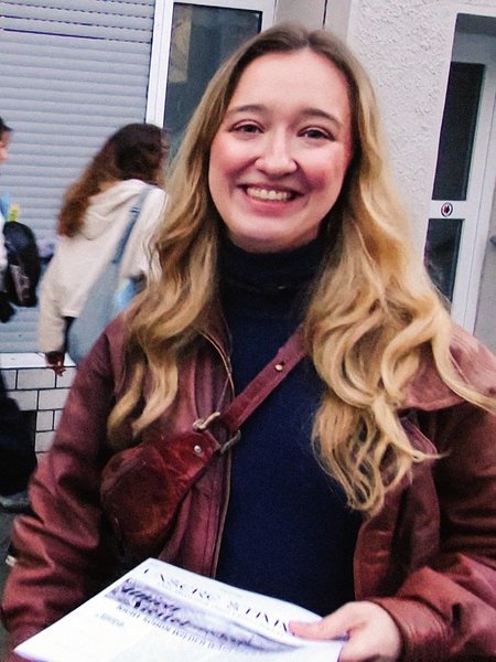 Eine junge Frau steht auf der Straße und lacht in die Kamera, sie hält einen Stapel Flugblätter in der Hand, im Hintergrund gehen Menschen vorbei.