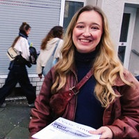 Eine junge Frau steht auf der Straße und lacht in die Kamera, sie hält einen Stapel Flugblätter in der Hand, im Hintergrund gehen Menschen vorbei.