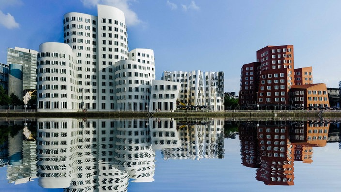 Drei verschiedene Gebäudekomplexe, eins weiß, eins glänzend silber und eins backsteinrot, stehen nebeneinander am Wasser, im Wasser spiegeln sich ihre Silouetten 