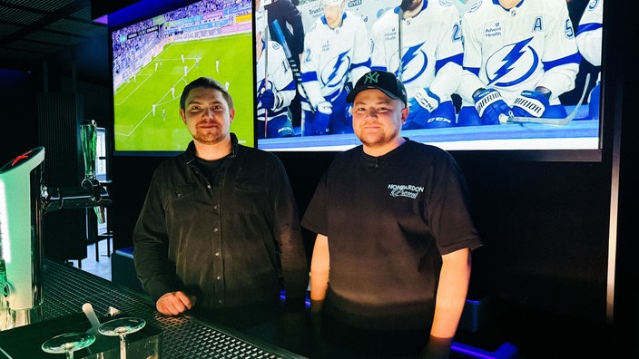 Roman und Kirill hinter der Theke der Youbar. Hinter ihnen hängen große Bildschrime an den Wänden, die Sport zeigen. 