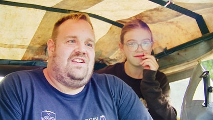 Florian Lindlein, ein blonder Mann im blauen T-Shirt, sitzt unter der hellen Plane eines Traktor-Führerhauses, rechts neben ihm sitzt ein junges Mädchen.