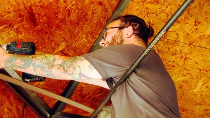 Ein braunhaariger, bärtiger Mann mit Brille hält in der rechten Hand eine Bohrmaschine, sein linker Arm ist frei und tätowiert. Über ihm ist eine Metallkonstruktion zu sehen, die mit Spanplatten bedeckt ist.