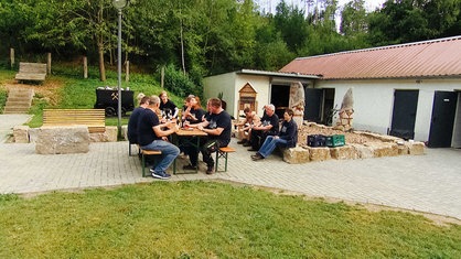 Mehrere Menschen sitzen auf Bänken und Steinen und essen gemeinsam.