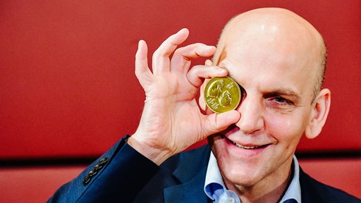 Benjamin List im Portrait. Er hält sich die Nobelpreismedaille vor das rechte Auge.