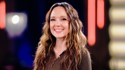 Carolin Kebekus, eine Frau mit gewellten langen braunen Haaren, trägt einen braunen Pullover und lächelt in die Kamera.