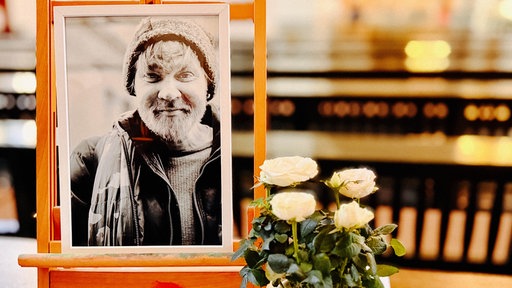 Ein Schwarz-Weiß-Foto in einem Rahmen, das Olli zeigt. Davor stehen weiße Rosen.