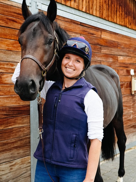Chantal Warmer trägt Reitkleidung und steht neben einem dunkelbraunen Pferd vor einem Stall.