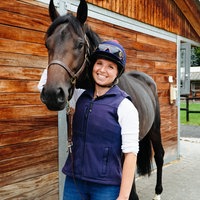 Chantal Warmer trägt Reitkleidung und steht neben einem dunkelbraunen Pferd vor einem Stall.