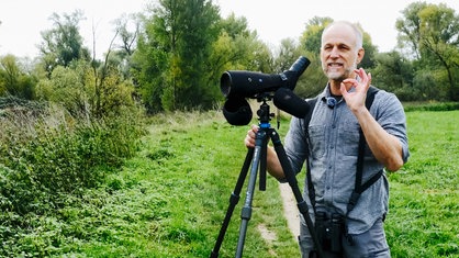 Ralph Achenbach steht auf einem Feldweg neben seinem Spektiv auf einem Stativ. Hinter ihm sind verschiedene Bäume und Sträucher.