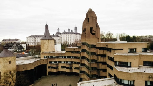Leicht welliges Gebäude aus braunem Beton mit unförmigem Turm, links mittelalterlicher Burgtürme, im Hintergrund ist ein weißes Schloss zu sehen
