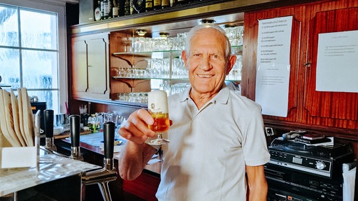 Toni Pinna, ein 86-jähriger Mann, hält ein Bier in der Hand und grinst.