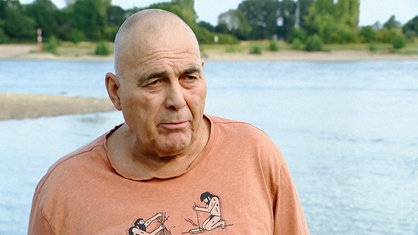 Jost Auler in einem orange-farbenen T-Shirt steht am Rhein.