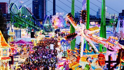 Blick in der Dämmerung auf eine Kirmes, mit bunt beleuchteten Fahrgeschäften und vielen Menschen