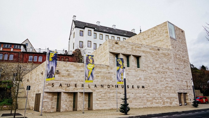 Neubau Sauerland-Museum in Arnsberg aus Sandstein, dahinter sieht man den mittelalterlichen Altbau, der über eine Brücke mit dem Neubau verbunden ist, vor dem Neubau wehen Fahnen, die die aktuelle Ausstellung bewerben