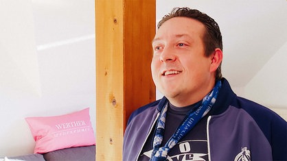 Lennart steht zuhause in seiner Wohnung. Er trägt eine Trainingsjacke und einen Schalke-Schal. Hinter ihm steht ein massiver Holzträger.