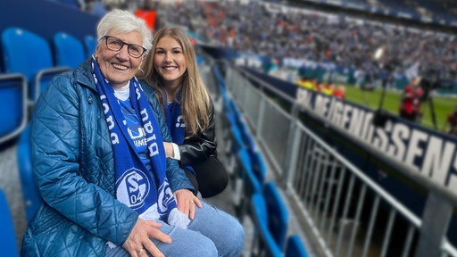 Rita und Theresa Stümper sitzen mit Fan-Schals um den Hals auf der Tribüne in der Schalke-Arena. Sie lächeln in die Kamera.