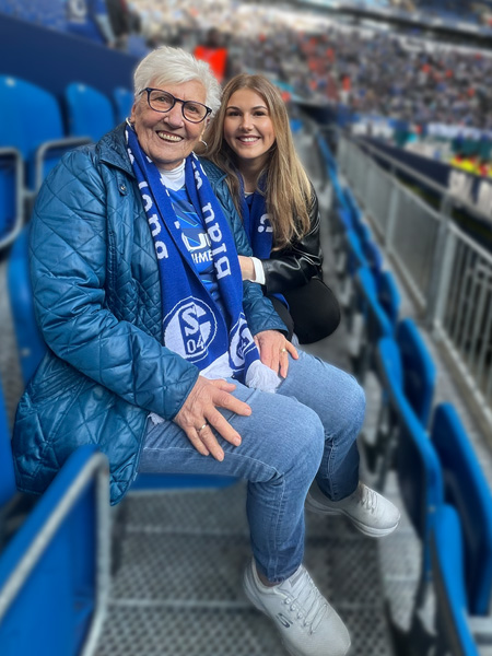 Rita und Theresa Stümper sitzen mit Fan-Schals um den Hals auf der Tribüne in der Schalke-Arena. Sie lächeln in die Kamera.