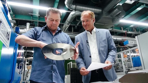 Eckard Jagalla steht in einer Produktionshalle. Er hält eine Schallplatte in den Händen und begutachtet sie. Rechts neben ihm steht der Geschäftsführer von Sonopress.
