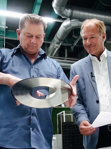 Eckard Jagalla steht in einer Produktionshalle. Er hält eine Schallplatte in den Händen und begutachtet sie. Rechts neben ihm steht der Geschäftsführer von Sonopress.