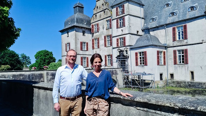 Ein Mann mit weißem Hemd und beiger Hose steht neben einer Frau mit blauem Hemd und beiger Hose, sie lehnen an einer Brücke, im Hintergrund sieht man ein Schloss samt Brücke aus Stein