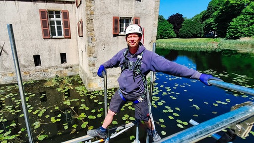 Mann steht in Arbeitskleidung, gesichert über einen Gurt, auf einem Gerüst über dem Wasser, im Hintergrund sieht man Burgmauern