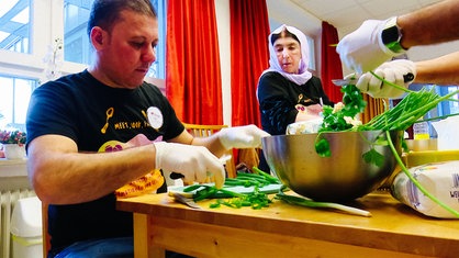 Eine ältere Frau mit weißem Kopftusch und ein mittelalter Mann mit schwarzen Haaren sitzen an einem hellbraunen Holztisch und schneiden Petersilie und Lauchzwiebeln