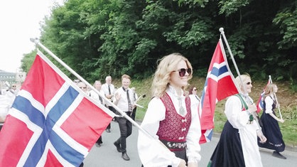 Beim Schützenfestumzug trägt Ana in ihrer Tracht eine norwegische Flagge. Mit ihr marschieren weitere Menschen, darunter auch Musiker. 