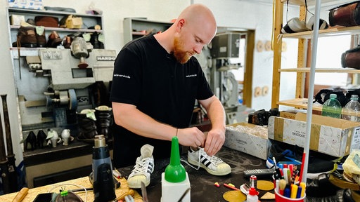 Patrick Rasche arbeitet an einem weißen Sneaker. Die Umgebung ist eine Werkstatt mit allerlei Materialien und Werkzeugen