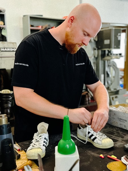Patrick Rasche arbeitet an einem weißen Sneaker. Die Umgebung ist eine Werkstatt mit allerlei Materialien und Werkzeugen