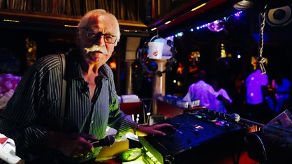 DJ Dietmar Duhme legt eine Hand auf sein Mischpult, in der anderen hält er ein Mikrofon. Im Hintergrund tanzen Menschen auf der Tanzfläche.
