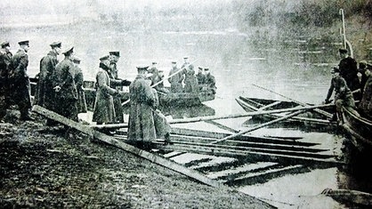 Ein Schwarz-Weiß-Bild, das Männer in Uniform an einem Flußufer zeigt, im Wasser schwimmen Holzstücke