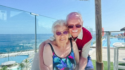 Regina Schmer, eine ältere Frau mit weißen Locken mit Sonnenbrille und Badeanzug, sitzt. Hinter ihr steht Rudi Vollak, ein älterer Mann mit Sonnenbrille, und umarmt die Frau. 