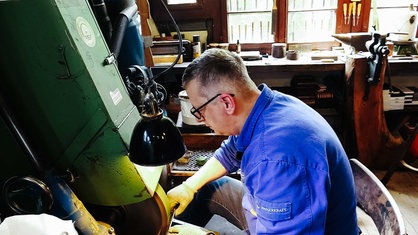 Mann mit blauem Hemd und Brille sitzt an einer Maschiene, er trägt Handschuhe und hält ein Messer an eine Schleife
