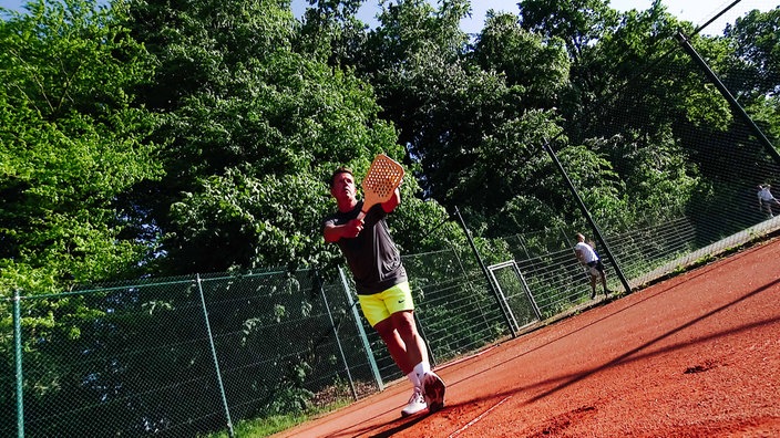 Ein Mann steht in Sportklamotten auf einem Sportplatz und hält einen Holzschläger und einen Ball vor sich.