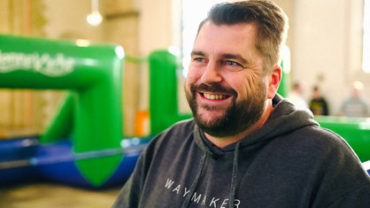Stefan Rapp steht in der Sportkirche und lächelt. Hinter ihm befindet sich das aufblasbare Fußballfeld.