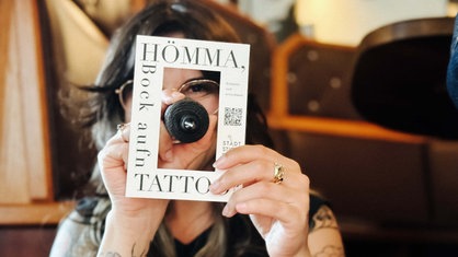 Melina Di Febo hält einen Postkarten-Rahmen und eine Tattoomaschine hoch. Sie schaut durch den Rahmen.