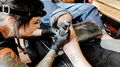 Melina Di Febo trägt eine Kopflampe. Sie sticht ein Tattoo auf der Wade von Rieke Sasse.