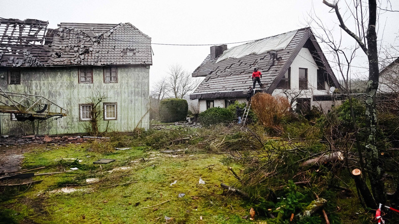 Zwei Häuse, beide sind zum Teil zerstört. Ihnen fehlen Dachpfannen. Die Wiese davor ist übersäht von Gestrüpp und umgeknickten Bäumen.