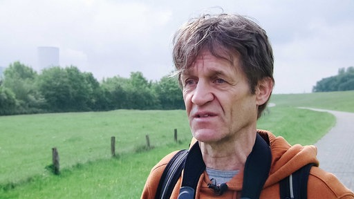 Ein Mann blickt nach vorne, er trägt einen orangenen Pullover, links im Hintergrund sieht man eine grüne Wiese