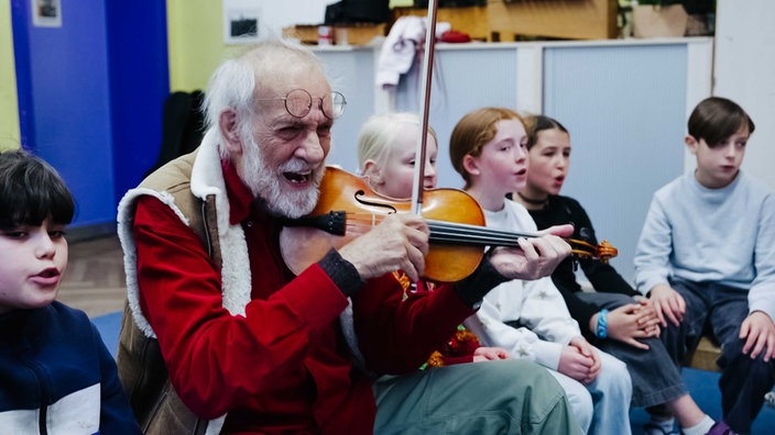 Mann mit weißem Bart und Brille auf der Stirn spielt Geige, um ihn herum sitzen Kinder und singen 