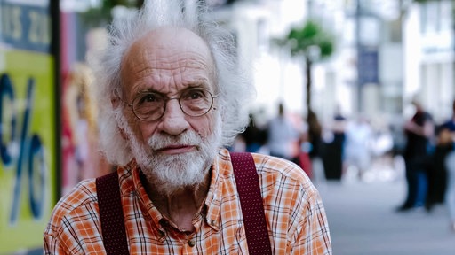 Mann mit weißem, schütteren Haar und Bart, runde Brille, orange-kariertes Hemd und roten Hosenträgern