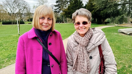 Zwei ältere Frauen stehen im Park nebeneinander und schauen in die Kamera. Die linke Frau trägt einen pinken Mantel und die rechte Frau eine graue Brille.
