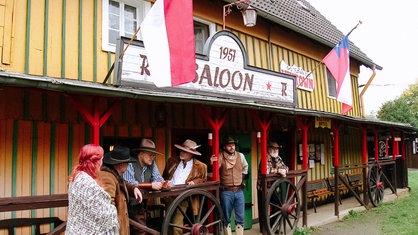Mehrere Männer und eine Frau stehen auf der Veranda eines "Saloons" und unterhalten sich.