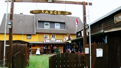 Im Vordergrund ein Holztor, auf dem "Ranch" steht. Dahinter steht ein gelbes Haus mit dem Schild "Saloon" über der Tür. Davor stehen mehrere Menschen in Westernkleidung.