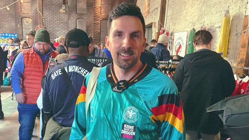 Christian Rottmeier, ein schwarzhaariger Mann mit Dreitagebart, steht im türkisen Trikot vor einer Kleiderstange in einer roten Backsteinhalle.