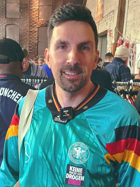 Christian Rottmeier, ein schwarzhaariger Mann mit Dreitagebart, steht im türkisen Trikot vor einer Kleiderstange in einer roten Backsteinhalle.