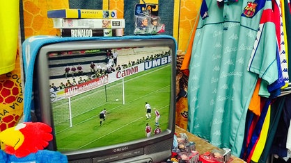 Auf einem alten Röhrenfernseher läuft ein Fußballspiel, daneben sieht man ein türkises Vintage-Fußballtrikot.