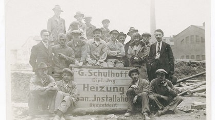 Viele Personen befinden sich in einer zerstörten Landschaft. Sie stehen um ein Schild. Darauf steht "G. Schulhoff Heizungen".