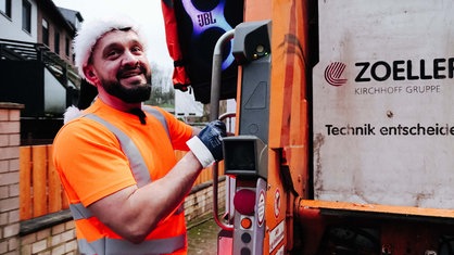 Alex Rubchev steht an einem Müllwagen. Auf dem Kopf trägt er eine Weihnachtsmann-Mütze. Er lächelt.